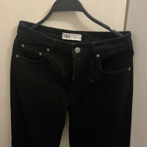 Svarta byxor med strass från Zara - Svarta byxor från Zara med snygga vågformade strassdetaljer på både fram- och baksida. Klassisk femficksmodell med knapp och dragkedja. Byxorna har en stilren look med en touch av glitter som gör dem extra unika.