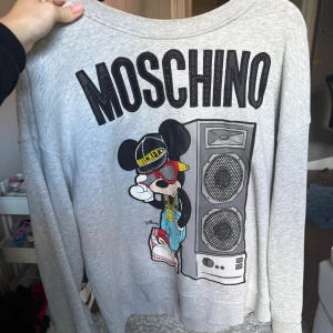 Grå Moschino tröja med Mickey Mouse - Snygg grå sweatshirt från Moschino med stort tryck av Mickey Mouse i streetstyle och högtalare på framsidan. Tröjan har rund hals, långa ärmar och är i mjuk bomullsmix. Perfekt statement piece för dig som gillar coola prints och designerplagg.