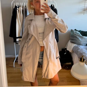 Beige trenchcoat med stora slag - Snygg beige trenchcoat med stora slag och dubbelknäppning. Jackan har långa ärmar, klassisk krage och bälte i midjan. Perfekt för lager-på-lager och ger en clean look till alla outfits.