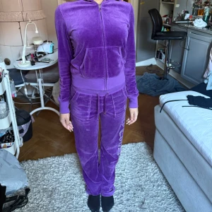 Lila Juicy Couture mjukisbyxor och hoodie - Säljer ett par lila mjukisbyxor samt hoodie från Juicy Couture i mjuk velour med glittrig logga längs benet. Byxorna har snörning i midjan, raka ben och fickor med lock bak. Perfekta för en chill och trendig look.