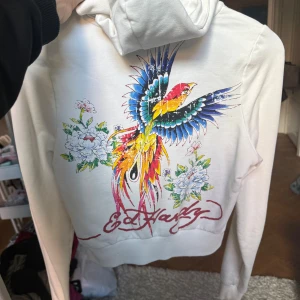 Vit Ed Hardy hoodie med färgglad fågel - Säljer en vit hoodie från Ed Hardy med dragkedja och huva. Baksidan har en stor, färgglad fågel och blommor i blått, gult och rött, samt Ed Hardy-logga. Framsidan har tryck med årtal och fickor. Perfekt statement-plagg för dig som gillar unika prints.