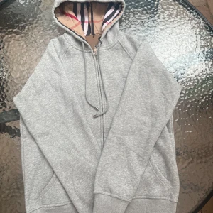 Grå Burberry hoodie med dragkedja - Snygg grå hoodie från Burberry med klassiskt rutigt foder i huvan och broderad logga på bröstet. Tröjan har dragkedja, snörning i huvan och kängurufickor. Perfekt för dig som vill ha en stilren och lyxig look med streetkänsla. Det är en XL men mycket liten i storleken. Den är som en M