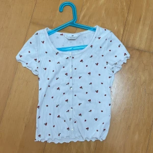 Vit topp med körsbär från H&M - Supersöt vit topp från H&M med små röda körsbärsmönster över hela. Toppen har korta ärmar, knappar framtill och vågig kant nertill och vid ärmslut. Perfekt för en somrig och lekfull stil!