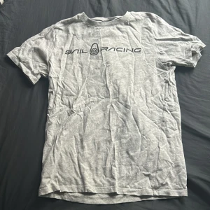 Grå t-shirt från Sail Racing - Snygg grå t-shirt från Sail Racing med svart logga tryckt över bröstet. Klassisk rund halsringning och korta ärmar. Tillverkad i mjuk bomull som känns skön mot huden. Perfekt för en avslappnad och sportig look.