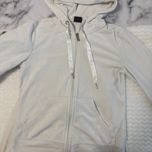 Vit velour mjukisdress med huva - Säljer en vit mjukisdress i velour med hoodie och matchande byxor. Hoodien har dragkedja, fickor och breda satinband i snörningen. Byxorna har resår i midjan och samma snygga satinband. Perfekt för chill dagar och riktigt skön mot huden. Kofta i storlek XS och byxorma i storlek S.
