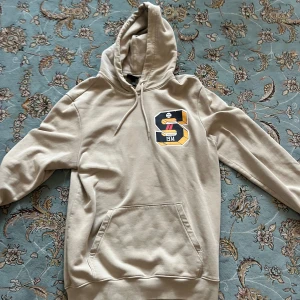 Beige hoodie med tryck från H&M - Säljer en beige hoodie från H&M i storlek M med stort S-tryck och baseballmotiv både fram och bak. Tröjan har huva med snörning, känguruficka och regular fit. Materialet är mjuk bomull och polyester, perfekt för chill dagar.