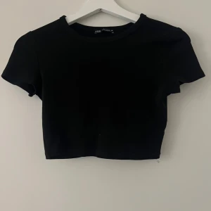 Svart croppad topp från Zara - Enkel och stilren svart croppad topp från Zara med rund halsringning och korta ärmar. Perfekt att matcha med höga jeans eller kjol för en clean look.
