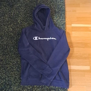 Mörkblå Champion hoodie 2XL - Snygg mörkblå hoodie från Champion i storlek 2XL. Klassisk modell med vit logga på bröstet, huva och känguruficka. Perfekt för dig som gillar streetwear och vill ha en bekväm och stilren tröja.