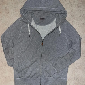 Grå hoodie med dragkedja från Morris - Snygg grå hoodie från Morris med vit snörning i huvan och dragkedja framtill. Tröjan har långa ärmar, två fickor och en liten broderad logga på bröstet. Perfekt för chill dagar eller när du vill vara bekväm.