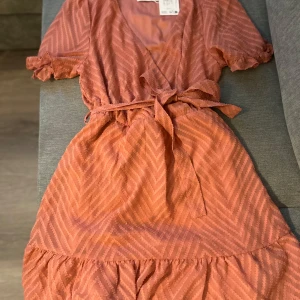 Kort omlottklänning från Vila, old rose - Superfin kort klänning från Vila i färgen old rose. Omlottmodell med knytband i midjan, volangkant nertill och korta ärmar med lätt rynk. Snyggt mönster i tyget som ger extra detalj. Perfekt för dig som gillar en feminin och trendig look. Aldrig använd