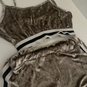 Beige sammetstopp med sportig kant - Supersnygg beige linnetopp och shorts i mjuk sammet med smala axelband. Toppen har en sportig känsla tack vare den vita och svarta randen nedtill. Perfekt för dig som gillar en chill men ändå trendig look.
