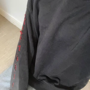 Svart hoodie med röd snörning  - Svart hoodie från lager 157 i storlek M med huva och långa ärmar. Unik detalj med röd snörning längs ena ärmen som avslutas med en rosett. Mjuk bomullskänsla och ribbade muddar vid ärmslut och nederkant. Perfekt för dig som gillar streetstyle och coola detaljer.