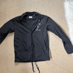 Svart vindjacka från Edwin - Svart vindjacka från Edwin med stor E-logga på bröstet. Jackan har krage, tryckknappar framtill, två fickor och snörning vid nederkanten. Lätt och smidig design i syntetmaterial, perfekt för dig som gillar streetwear och enkel stil.