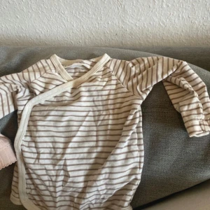 Beige och vit randig baby body 1-3 mån - Säljer en beige och vit randig omlotttröja med långa ärmar. Tröjan har praktiska tryckknappar längs sidan och är gjord i mjuk bomull, perfekt för chill dagar. Enkel och stilren design som passar till allt.