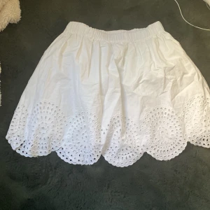 Vit kortkjol med broderad spetskant - Supersöt vit kortkjol från C i storlek XS. Kjolen har en elastisk midja och en broderad spetskant med snygga hålmönster längs nederdelen. Perfekt för sommarens alla tillfällen när du vill känna dig extra fräsch och stylish.