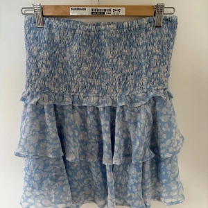 Blå och vit-mönstrad volangkjol från Lindex - Supersöt kort kjol från Lindex i ljusblått med vitt mönster. Kjolen har smock upptill och volanger nedtill. Kan även användas som topp! Använd typ 3 gånger så den är som ny! Storlek S. Säljer då den inte kommer till användning.