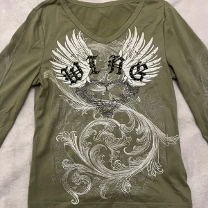 Olivgrön långärmad tröja med tryck  - Säljer en fin olivgrön långärmad tröja med coolt vingmotiv och text. Mycket bra skick, säljer pågrund att det inte är min stil. Köpt från plick men märken är shein. Fråga om ni undrar något ^^