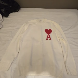 Vit stickad tröja från Ami Paris - Vit stickad tröja från Ami Paris med röd hjärtlogga och bokstaven A på bröstet. Klassisk rund halsringning och ribbade muddar. Perfekt för dig som gillar stilrena plagg med en twist. Säljer då den var för stor för mig. Använd en gång, inga defekter. Har inge kvitto då jag fick den i gåva.