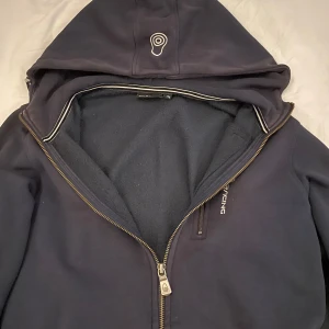 Mörkblå hoodie från Sail Racing - Mörkblå hoodie från Sail Racing med dragkedja och broderad logga på bröstet och huvan. Tröjan har huva med dragsko, lång ärm och mjukt fleecefoder på insidan. Perfekt för kyliga dagar och snygg till jeans eller joggers.