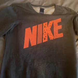 Grå Nike sweatshirt med orange tryck - Snygg grå sweatshirt från Nike med stort orange logotyp-tryck på bröstet. Tröjan har klassisk passform och rund halsringning, perfekt för en sportig och avslappnad stil. Materialet känns mjukt och bekvämt mot huden.