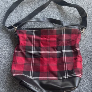 Rutig röd och svart axelväska - Säljer en stor axelväska med röd och svart rutig design. Väskan har bred axelrem i textil, detaljer och botten i svart skinnimitation samt dragkedja upptill. Insidan har flera fack med dragkedja för extra förvaring. Perfekt för dig som gillar en edgy och praktisk look.