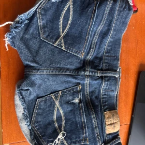 Mörkblå jeansshorts Abercrombie - Snygga mörkblå jeansshorts från Abercrombie & Fitch med fransiga kanter och slitna detaljer. Klassisk femficksmodell med dekorativa sömmar på bakfickorna. Perfekta för sommardagar och festivalhäng.