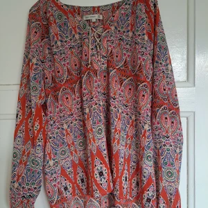 Mönstrad blus från Flash Woman L - Färgstark blus med paisleyinspirerat mönster i rött, rosa, grönt och blått. Blusen har långa ärmar, snörning vid halsen och en luftig passform. Perfekt för dig som gillar boho-vibbar och vill sticka ut med coola detaljer.