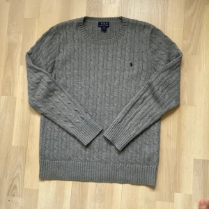 Grå kabelstickad tröja Polo Ralph Lauren - Kabelstickad grå tröja från Polo Ralph Lauren i storlek XL. Klassisk rund halsringning, långärmad och med broderad marinblå logga på bröstet. Perfekt för dig som gillar stilren och tidlös design med en preppy touch.