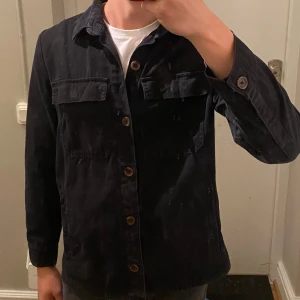 Svart overshirt  - Svart overshirt från Threadbare i bomull. Klassisk design med krage, bröstfickor med lock och knäppning med bruna knappar.