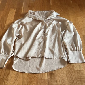 Beige skjorta från L&S med knappar - Stilren beige skjorta från L&S i polyester med klassisk krage och knäppning framtill. Skjortan har långa ärmar med breda manschetter och en något längre, rundad nederkant bak. Perfekt för dig som gillar en clean och enkel look.