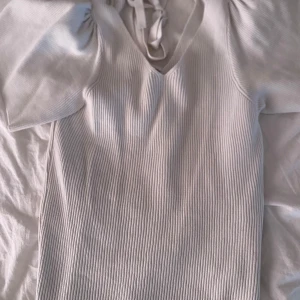 Vit ribbad topp med knyt från ONLY - Supersnygg vit ribbad topp från ONLY med v-ringning och knyt i nacken. Toppen har korta puffärmar och är tillverkad i ett mjukt, b material som sitter skönt. Perfekt för dig som gillar en stilren look med det lilla extra.
