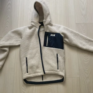 Beige teddyjacka Helly Hansen 140/10 - Mysig beige teddyjacka från Helly Hansen med huva och marinblå detaljer. Jackan har dragkedja framtill, två sidofickor och en bröstficka med HH-logga. 