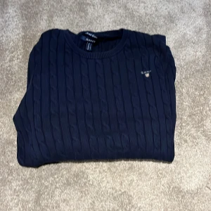 Marinblå kabelstickad tröja GANT - Snygg marinblå kabelstickad tröja från GANT i bomull. Klassisk rund halsringning och diskret logga broderad på bröstet. Perfekt för dig som gillar en stilren och tidlös look med lite preppy känsla. Den är i storlek L men väldigt liten i strl så skulle säga att den passar dig som bär mellan xs och S i vanlig fall