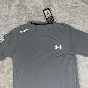 Grå Under Armour tränings-tshirt - Snygg grå t-shirt från Under Armour med UA-SPEC-tryck på axeln och klassisk logga på bröstet. Tillverkad i lätt och snabbtorkande polyester, perfekt för löpning eller gym. Rund halsringning och korta ärmar ger en sportig vibe.