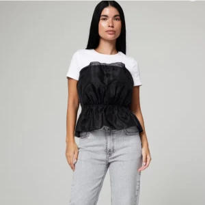 Svart bandeau topp med volang - Säljer en svart bandeau topp med markerad midja och volang nertill. Toppen har en glansig finish och är ärmlös, perfekt att styla över en t-shirt eller bära som den är. Snygg och trendig detalj till din outfit.( Prislapp är kvar, aldrig använd)