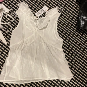 Vit volangblus med snörning - Supersöt vit blus från Kappahl med volangdetaljer vid halsringningen och snörning framtill. Blusen är ärmlös och har en lätt, luftig känsla med plisserad effekt i tyget. Perfekt för en fräsch och somrig look.