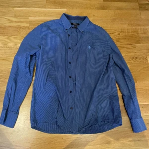 Blå randig skjorta - Snygg blå skjorta med tunna vita ränder och klassisk krage. Skjortan har svarta knappar, bröstficka med broderad logga och långa ärmar med manschett. Perfekt för dig som gillar stilren och enkel design.