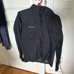 Svart vindjacka från Columbia - Svart vindjacka från Columbia med huva och dragkedja framtill, långa ärmar och justerbar huva.