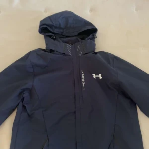 Mörkblå vindjacka från Under Armour - Säljer en mörkblå vindjacka från Under Armour med huva och dragkedja. Jackan har en sportig look med logga på bröstet och praktiska fickor. Perfekt för blåsiga dagar och utomhusaktiviteter. Men vill bara säga att den är liten i storleken. Den är storlek XL men den sitter som storlek M