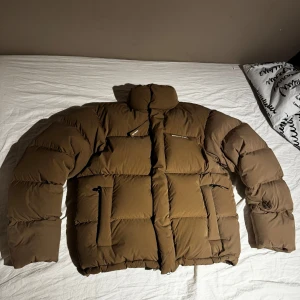 Brun pufferjacka från Axel Arigato - Säljer en brun pufferjacka från Axel Arigato med hög krage och dragkedja framtill. Jackan har två framfickor med dragkedja och tryckt logga på bröstet. Perfekt för kalla dagar när du vill hålla dig varm och snygg. Använd minimalt, enda tecken på användning är mitt namn på lappen, går att stryka över med vit penna sedan skriva nytt.