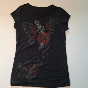 Svart t-shirt med strasstenar - Svart t-shirt med färgglatt motiv framtill dekorerat med strasstenar i rött, blått och grönt. T-shirten har rund halsringning och korta ärmar. Skriv gärna vid funderingar😊