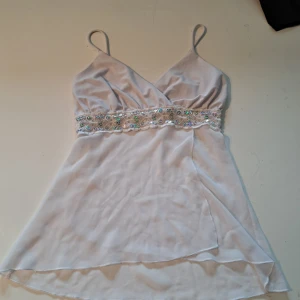 Vit topp med paljettdekor - Säljer en vit topp med smala axelband och omlott framtill. Toppen har en bred midjedetalj med glittrande paljetter. Tunt, lätt och transparent material som ger en luftig känsla. Perfekt för sommarens tillfällen! 