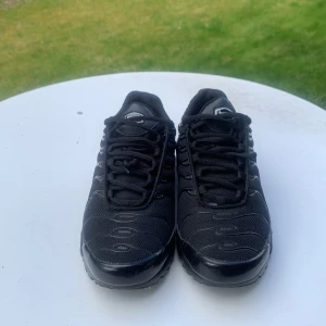 Svarta Nike Air Max Plus sneakers - Säljer ett par helsvarta Nike Air Max Plus sneakers med mesh- och syntetöverdel, tydliga vågformade detaljer och synlig Air-dämpning i sulan. Klassisk Nike-logga på sidan och plös. Perfekta för dig som gillar sportig stil.