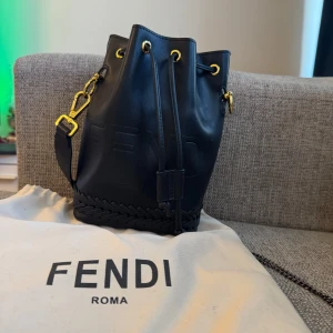 Svart bucket bag från Fendi - Säljer en svart bucket bag från Fendi med präglad logga och snygga guldfärgade metalldetaljer. Väskan har dragskostängning och flätad detalj längst ner. Insidan är fodrad i beige och har märkning 'Fendi Made in Italy'. Kommer med påsen som man ser på bild numer 5. Bara att skriva vid frågor och priset kan såklart diskuteras!