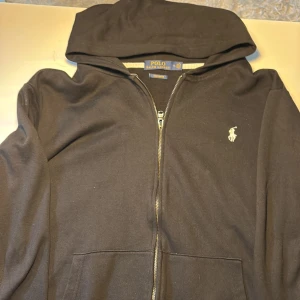 Svart hoodie från Polo Ralph Lauren - Säljer en stilren svart hoodie från Polo Ralph Lauren med dragkedja och det klassiska broderade logotypen på bröstet. Hoodien har en bekväm passform och är perfekt för en avslappnad look. Köp direkt eller ge prisförslag 💛