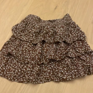 Brun volangkjol med blommigt mönster från Lindex - Supersöt brun kjol från Lindex med små vita blommor och flera volanger. Kjolen har resår i midjan och är perfekt för dig som gillar en lekfull och trendig stil. Lätt och luftig modell som passar till många olika outfits. Jättegullig till i sommar men även till någon annan årstid! Inte använd mycket alls! Jag passar i den och har storleken 158/164 men jag tycker att den passar 146/152 också! Skriv för bilder när jag har på mig den! Först till kvarn. 