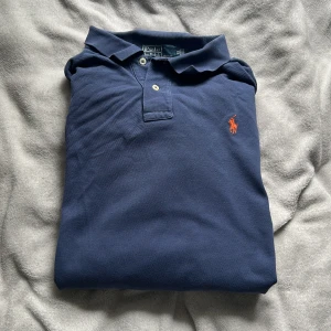 Blå långärmad pikétröja från Polo Ralph Lauren - Säljer en klassisk blå långärmad pikétröja från Polo Ralph Lauren med orange broderad logga på bröstet. Tröjan har krage och knappar framtill, perfekt för en stilren och avslappnad look.