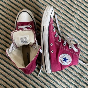 Mörkrosa Converse All Star sneakers - Säljer ett par klassiska mörkrosa Converse All Star high-top sneakers med vita snören och gummisula. Skorna har den ikoniska loggan på sidan och vita detaljer runt sulan och tån. Perfekta för dig som gillar en tidlös och cool stil.