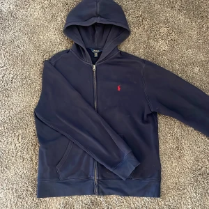 Mörkblå hoodie från Polo Ralph Lauren - Säljer en mörkblå hoodie från Polo Ralph Lauren med dragkedja och huva. Tröjan har en röd broderad logga på bröstet och två fickor framtill. Perfekt för en avslappnad stil. Storleken är i barn så skulle passa XS möjligtvis S. Hör av vid funderingar!
