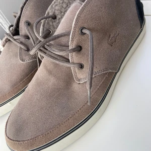 Beige mockasneakers från Lacoste - Säljer ett par snygga beige sneakers från Lacoste i mocka med snörning och vit platt sula. Skorna har rund tå och diskret Lacoste-logga på sidan. Perfekta för dig som gillar stilrena och bekväma skor.